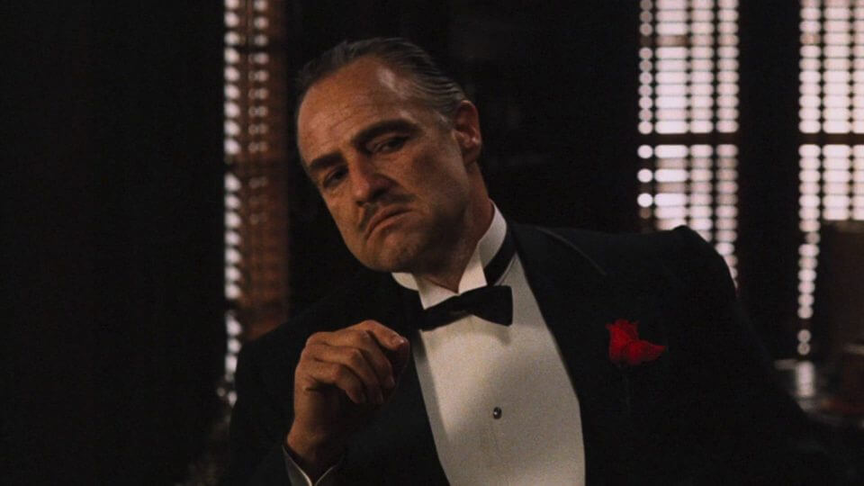 Don Vito Corleone, patriarca da família Corleone, de terno preto em ambiente escuro